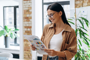 Charming caucasian woman wearing glasses holding papers and looking at it while standing at the office. Business lady with documents