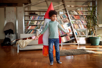 A little boy with a rocket toy on his back is posing at home while imagining flying to the space. Home, game, childhood