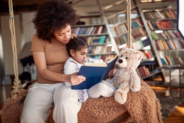 A young mother is reading a book to her little daughter while sitting on the swing at home together. Family, together, leisure