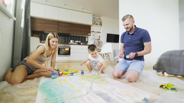 joyful family surrounds large handmade picture and delighted little boy throws paintbrush on canvas near smiling mother painting blue sky at home