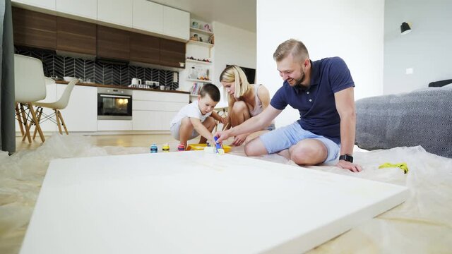 joyful family surrounds large handmade picture on floor at home. Mother father and little preschool son spending time together in modern apartment, drawing big picture, low angle view