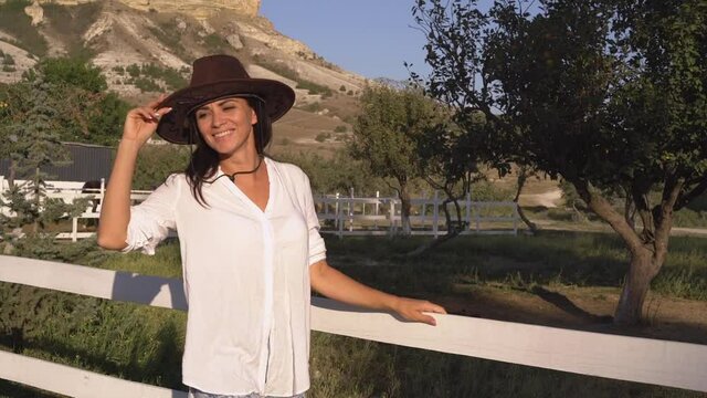 Young beautiful woman in hipster hat and white shirt posing outside in green country ranch near fence with garden on summer mountain background on sunny day, people lifestyle concept