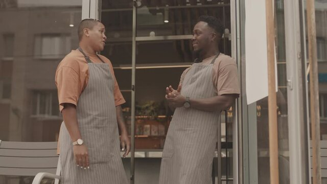 Medium long of young Black male and Biracial female waiters wearing aprons, standing at door outside, chatting and smiling