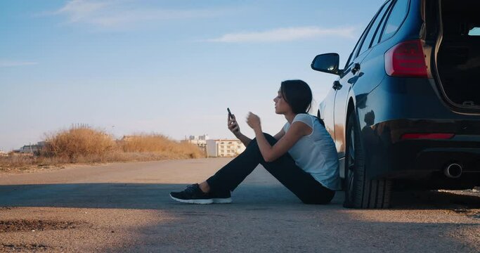 Sad and frustrated beautiful woman sitting a side of vehicle with flat rear tire on country road during travel and using smartphone to find the service or calling to insurance for tow truck 