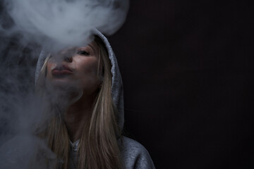 Portrait of beautiful blonde girl in gray hoodie smokes vape isolated on black studio background, cloud of steam smoke, mini hookah