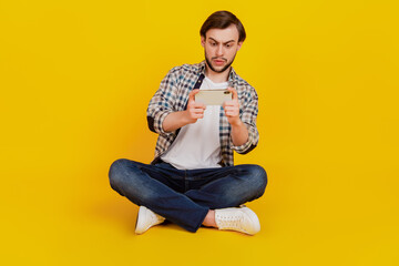 Full length photo of positive cheerful man sit floor play game cellphone isolated yellow color background