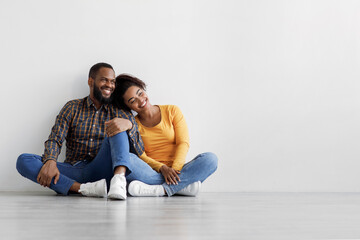 Satisfied young african american male and female sitting on floor on gray wall background, hugging, planning interior