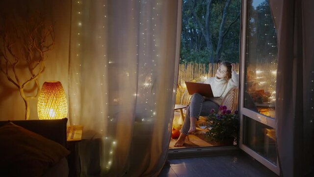 Curly haired young woman wearing warm sweater types on laptop sitting on balcony decorated with lights and autumn attributes against dark trees outside