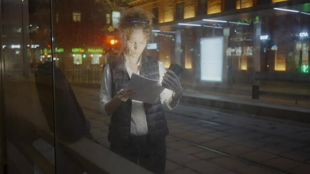 Office worker woman with short haircut stands on transport stop and looks at documents highlighting with smartphone against tram in late evening