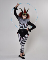 Full length  portrait of red haired  girl wearing a black and white clown jester costume, theatrical circus character.  Standing pose with hula hoop, isolated on  studio background.
