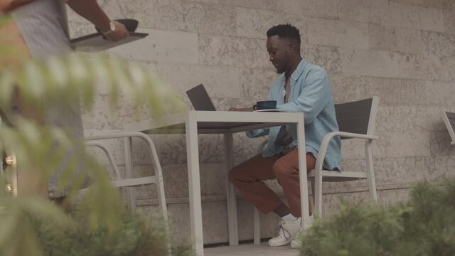 Low angle locked-down of cropped waiter serving lunch on tray to young Black man who sitting at table outdoors, using portable computer