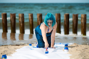 Talented blue-haired woman performance artist smeared with gouache paints on large canvas on beach
