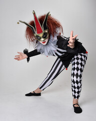 Full length  portrait of red haired  girl wearing a black and white clown jester costume, theatrical circus character.  Sitting down on floor, isolated on  studio background. - obrazy, fototapety, plakaty