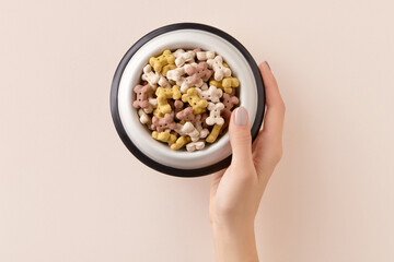 Womans hand holding dry dog food in bowl on beige background. Healthy organic nutrition for pets