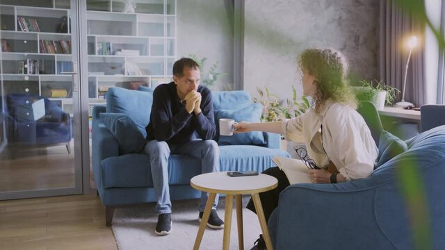 Curly haired woman consultant offers cup of coffee to stressed man client at appointment in stylish company office with stylish comfortable furniture
