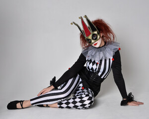 Fototapeta na wymiar Full length portrait of red haired girl wearing a black and white clown jester costume, theatrical circus character. Sitting down on floor, isolated on studio background.