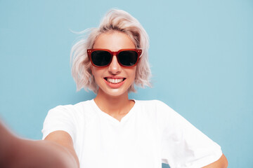 Young beautiful smiling female in trendy summer clothes. Sexy carefree woman posing near blue wall in studio. Positive blond model having fun in sunglasses. Cheerful and happy. Taking Pov selfie