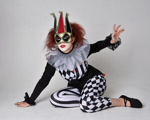 Full length  portrait of red haired  girl wearing a black and white clown jester costume, theatrical circus character.  Sitting down on floor, isolated on  studio background.