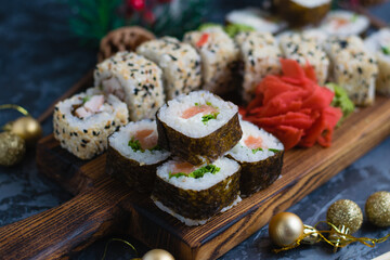 Large sushi set on a wooden board. Rolls with sesame seeds, nori, cheese, red fish, cucumber and eel, ginger, wasabi and christmas decor on gray background
