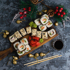 Large set of sushi on a wooden board top view. Rolls with sesame seeds, nori, cheese, red fish, cucumber and eel, ginger, wasabi, soy sauce, bamboo sticks and christmas decor on gray background