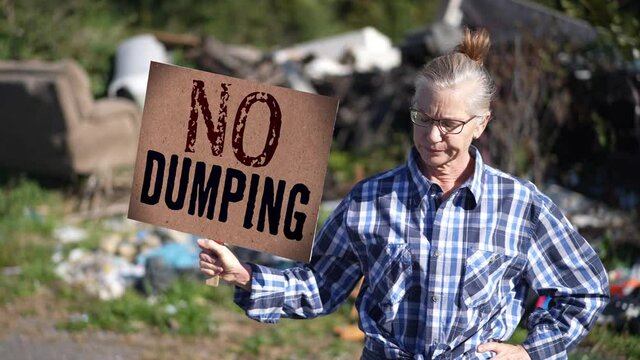 Unhappy, sad woman holding No Dumping sign in front of dumped garbage on sunny day.