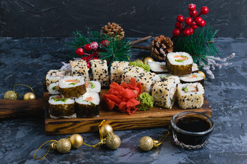 Rolls with sesame seeds, nori, cheese, red fish, cucumber and eel, ginger, wasabi, soy sauce and Christmas decor on a gray background. A large set of sushi on a wooden board.