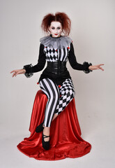 Full length  portrait of red haired  girl wearing a black and white clown jester costume, theatrical circus character.  Sitting down on chair, isolated on  studio background.