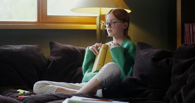 A girl in glasses reading a book with a dreamy look, sitting on the sofa