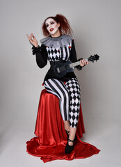 Full length  portrait of red haired  girl wearing a black and white clown jester costume, theatrical circus character.  Sitting down on chair, isolated on  studio background.