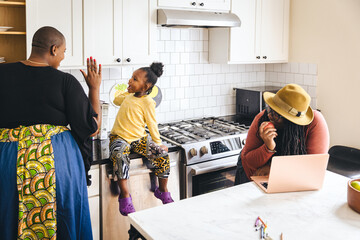 Mother high-fives her daughter in the kitchen while the other parent works on a laptop