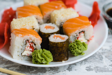 Sushi on a white plate on a light background with copy space. Delicious rolls with cheese, fish, sesame seeds, crab stick, wasabi, ginger and soy sauce.