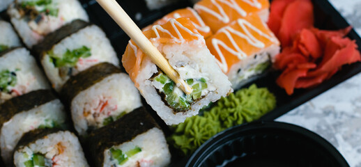 Hand holds wooden sticks sushi california roll with fish, cheese and mayonnaise. Large set of rolls with soy sauce and wasabi on the background