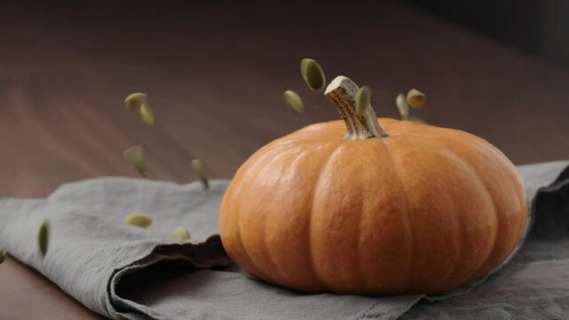 Slow motion pumpkin seeds fall on orange pumpkin on walnut table with linen cloth with copy space