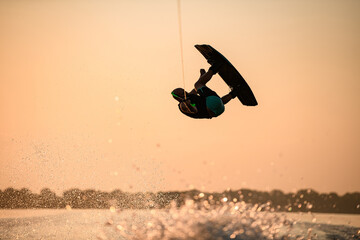 man making trick in jump with wakeboard on bright orange sky and sun background.