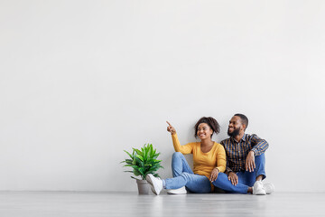 Dreaming smiling african american couple sitting on floor with plant in pot on gray wall background