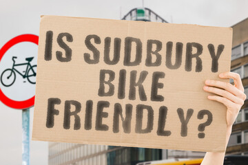 The question " Is Sudbury bike friendly? " on a banner in men's hand with blurred background. Transportation. Zero waste. Bicycle lane. Streets. City. Safety. Insecure. Road signs. Dangerous