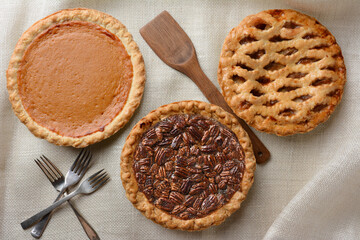 Three fresh baked holiday pies, Apple, Pecan and Pumpkin on a burlap table cloth. Horizontal format with forks and wooden spatula. © Steve Cukrov