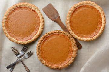 High angle shot of three fresh baked Thanksgiving Pumpkin pies, on a burlap table cloth. Horizontal format with forks and wooden spatula.
