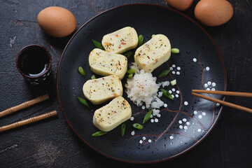 Tamagoyaki or sweetened japanese egg roll served with daikon and green onion on a black plate, studio shot on a dark brown stone surface