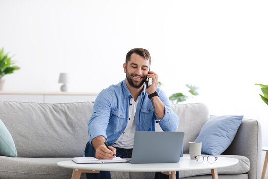 Smiling adult european man with beard sits on sofa work at home remotely with laptop, talk to client by phone