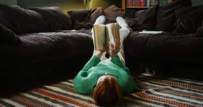 A girl reading a book with lying on the floor and resting legs on sofa