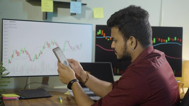 Said view of Trader at home office watching stocks on mobile phone while multiple graphs on monitors - concept of investing in cryptocurrency on smartphone app, studying share market and trading.