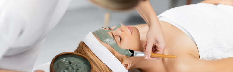 beautician holding bowl and applying clay mask on face of woman in spa center, banner