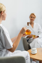 blonde spa specialist holding glass jug with massage oil near blurred client