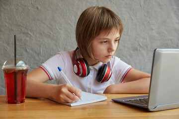 schoolgirl with headphones sits at table, watching at laptop screen, writing notes at copybook. little caucasian girl studying with computer, doing homework. distant learning, online education, lesson