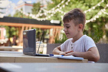 little schoolboy sits outdoors with laptop. smiling caucasian boy sitting at table at veranda of cafe, watching digital lesson at screen of computer, doing homework. distant learning, online education