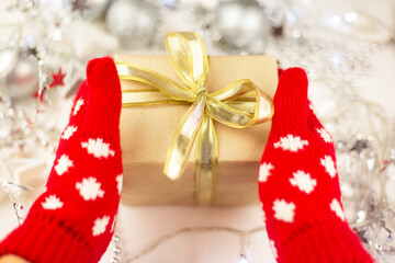 Female hands hold a Christmas gift with a gold ribbon in red mittens, new year mood, preparation for the celebration.