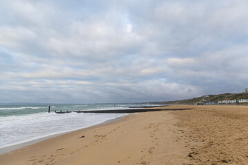 A scenic view of a sandy beach with crashing waves, wooden groynes and some grassy dunes in the background under a grey stormy sky