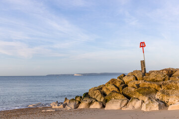 A scenic view of a rocky groyne with a red wind post along a sandy beach with a majestic blue sea and white hilly cliffs in the background under a majestic blue sky and some white clouds