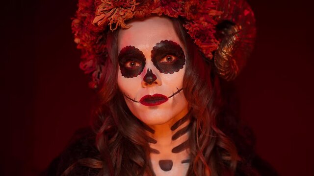 Caucasian female brunette in a Halloween image of a witch with the makeup of a painted skull with ram horns, sewn her mouth, flowers looking into the camera in a dark fiery room close-up